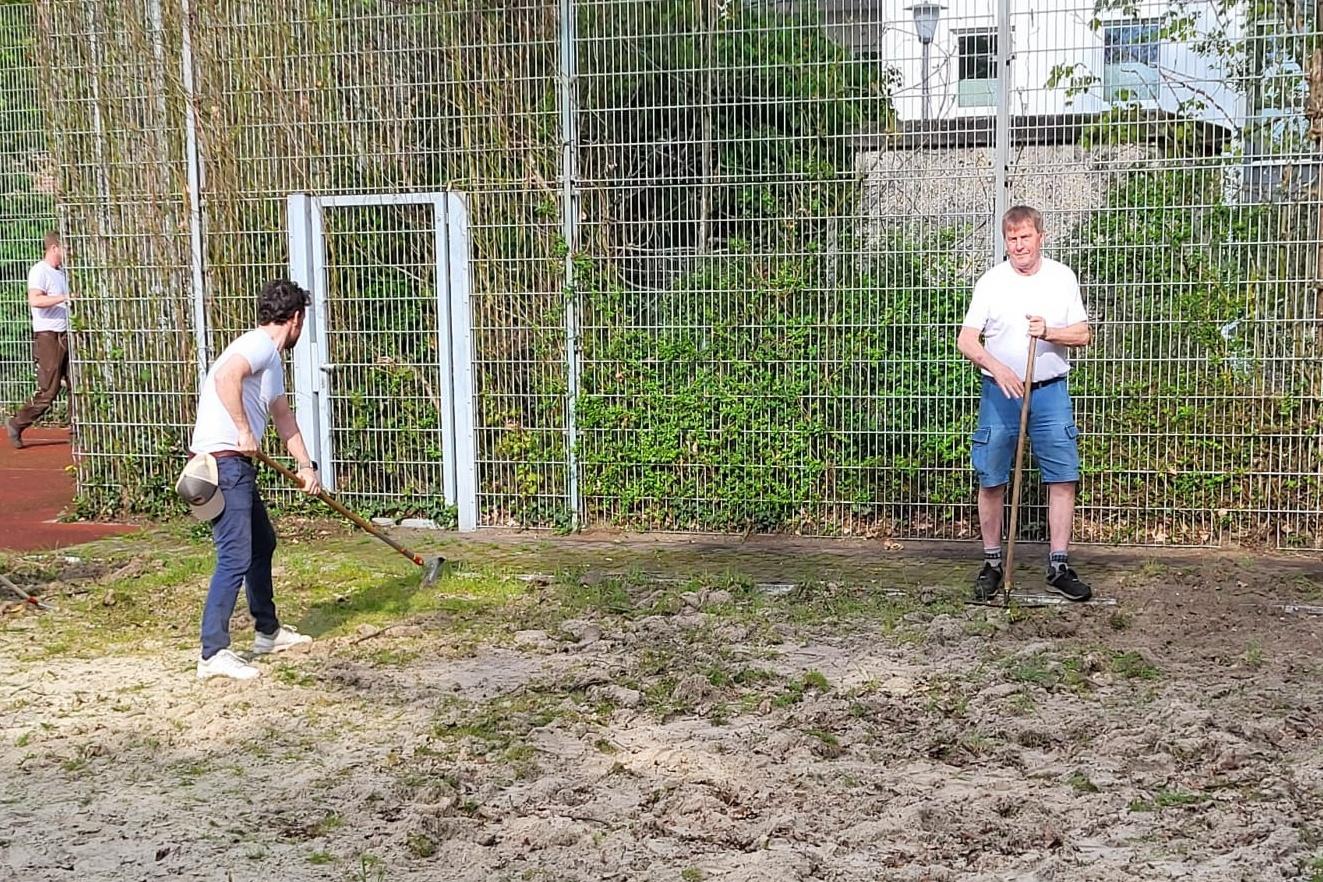 Drei Personen arbeiten mit Schaufeln auf einem sandigen Sportplatz hinter einem Fu&szlig;balltor.