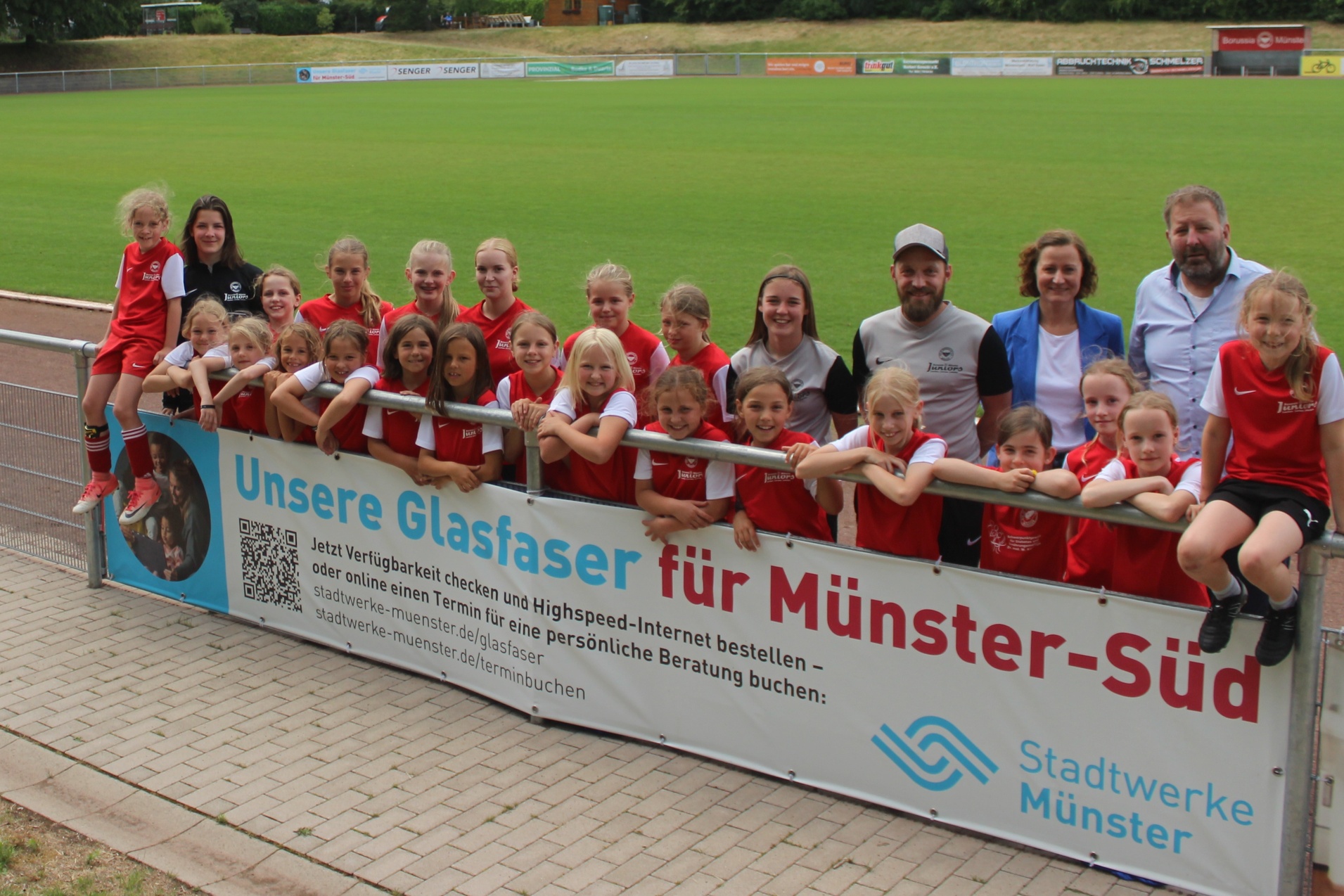 Gruppenshot von M&auml;dchenmannschaft in roten Trikots, posierend am Zaun eines Fu&szlig;ballfeldes mit einem Werbebanner.