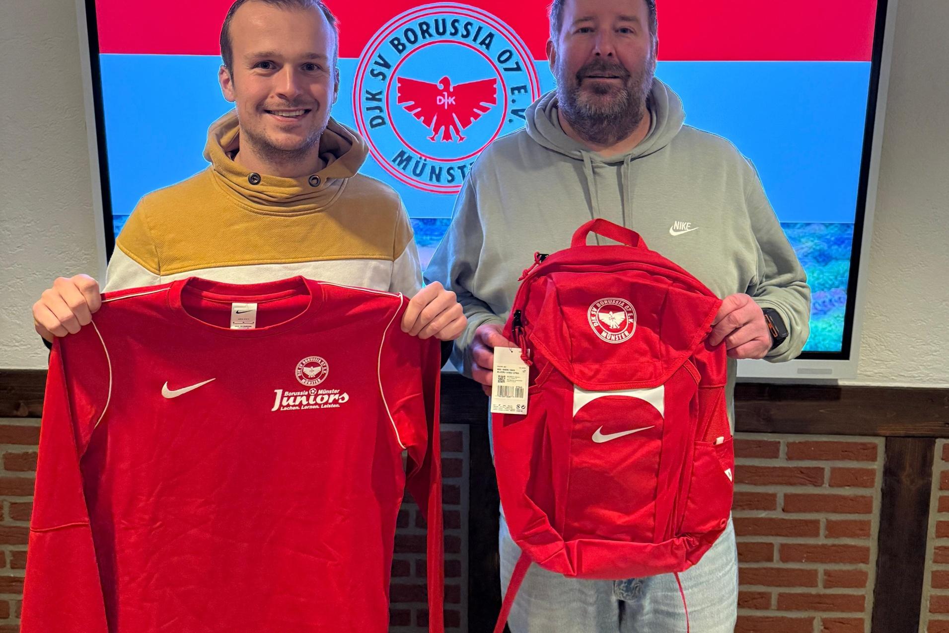Zwei M&auml;nner halten ein rotes T-Shirt und einen roten Rucksack vor einem Bildschirm mit dem Logo von Borussia M&uuml;nster.