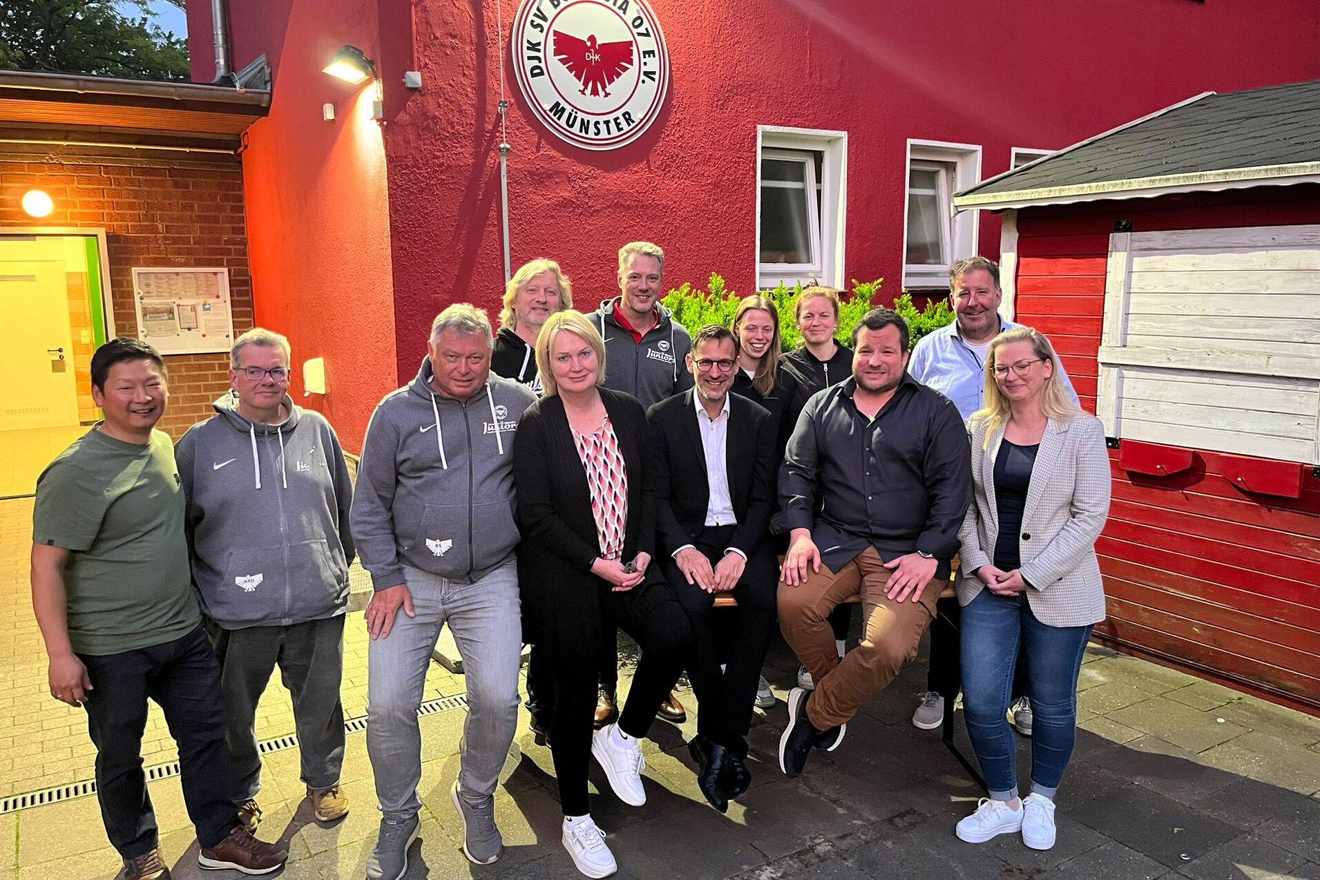 Gruppenzusammenkunft vor der Vereinsfassade von Borussia M&uuml;nster, Mitglieder posieren l&auml;chelnd in zwei Reihen.
