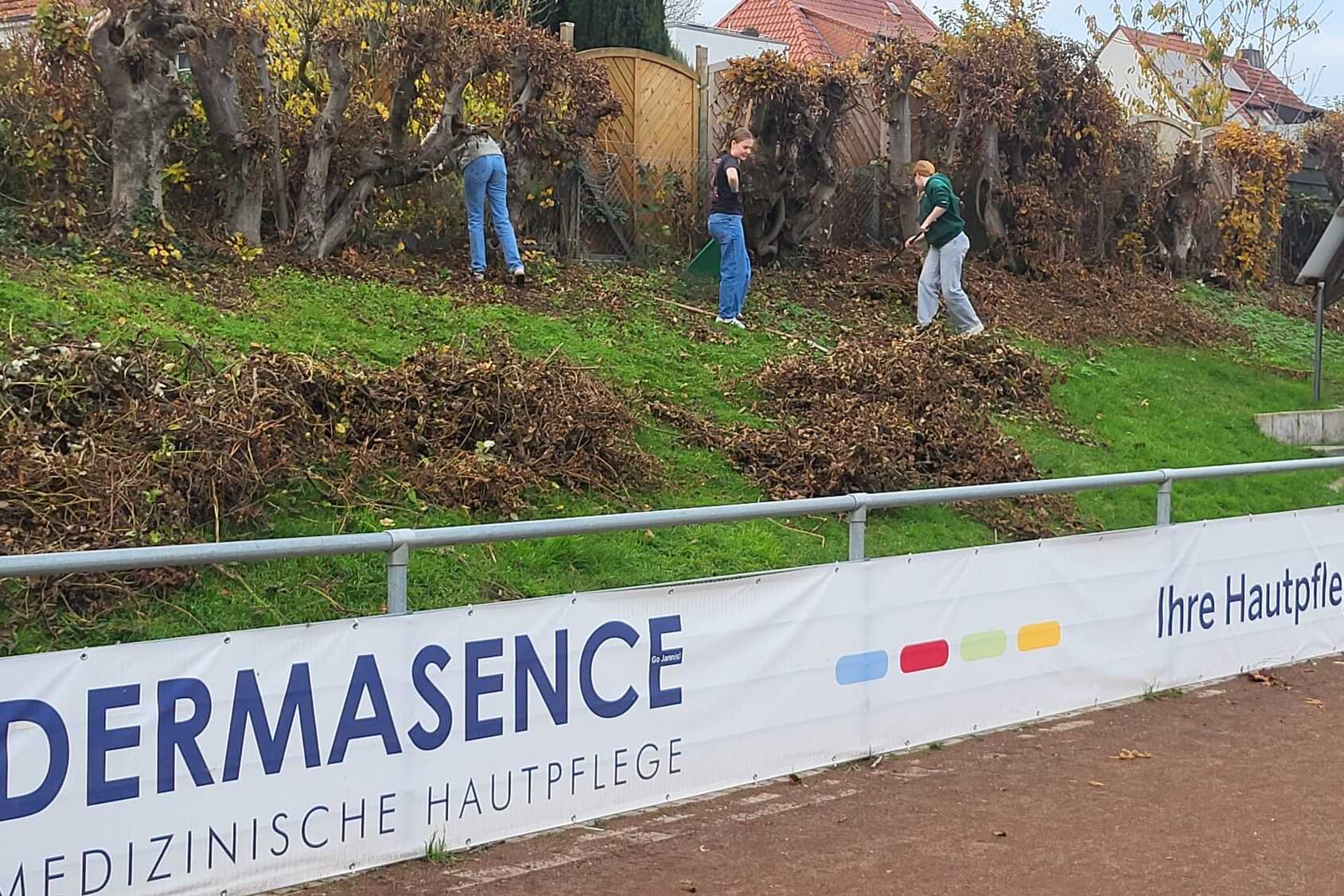 Drei Personen entfernen Laub auf einem Hang, w&auml;hrend ein Werbebanner f&uuml;r Dermasence im Vordergrund sichtbar ist.