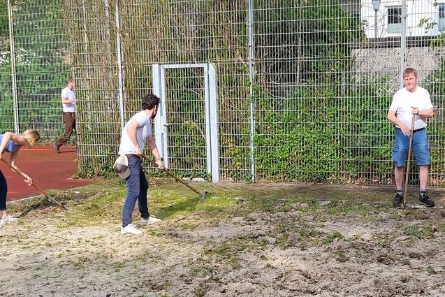 Drei Personen arbeiten mit Schaufeln auf einem sandigen Sportplatz hinter einem Fu&szlig;balltor.