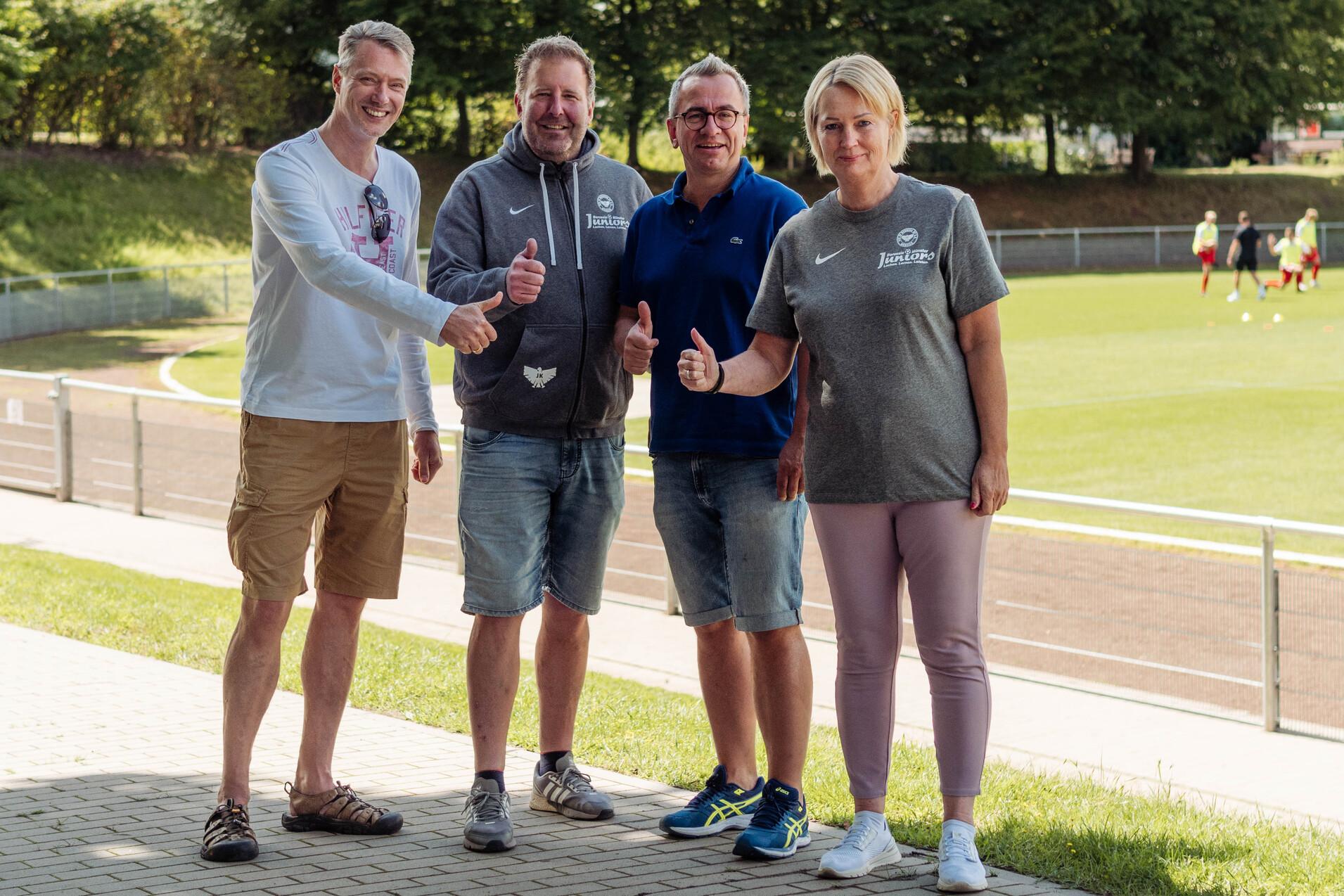 Vier Personen stehen l&auml;chelnd auf einem Sportgel&auml;nde und zeigen Daumen hoch, im Hintergrund Trainierende auf dem Platz.