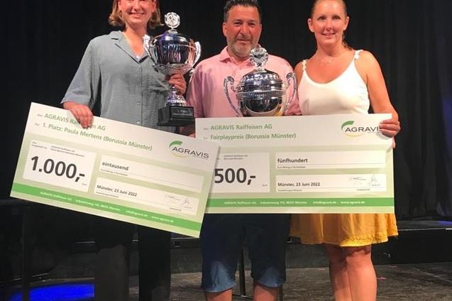 Drei Personen halten Pokale und Schecks f&uuml;r die Platzierungen bei einer Sportveranstaltung auf einer B&uuml;hne.