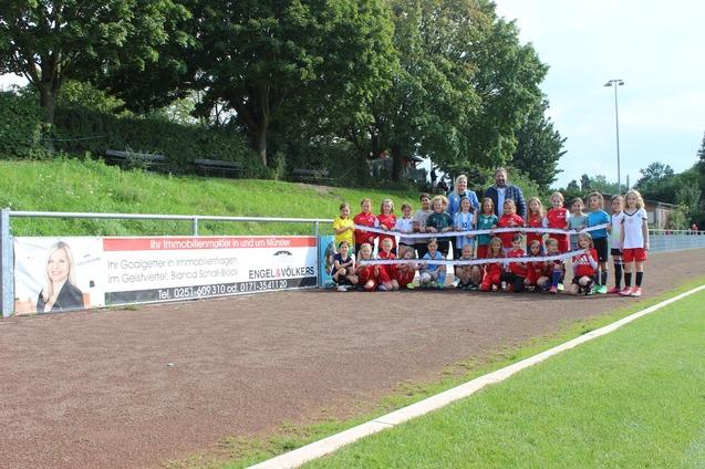 Gruppenfoto von jungen Fu&szlig;ballspielern in Vereinskleidung, posierend vor einem Werbebanner auf dem Sportplatz.