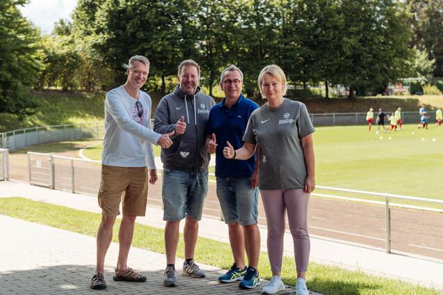 Vier Personen stehen l&auml;chelnd auf einem Sportgel&auml;nde und zeigen Daumen hoch, im Hintergrund Trainierende auf dem Platz.