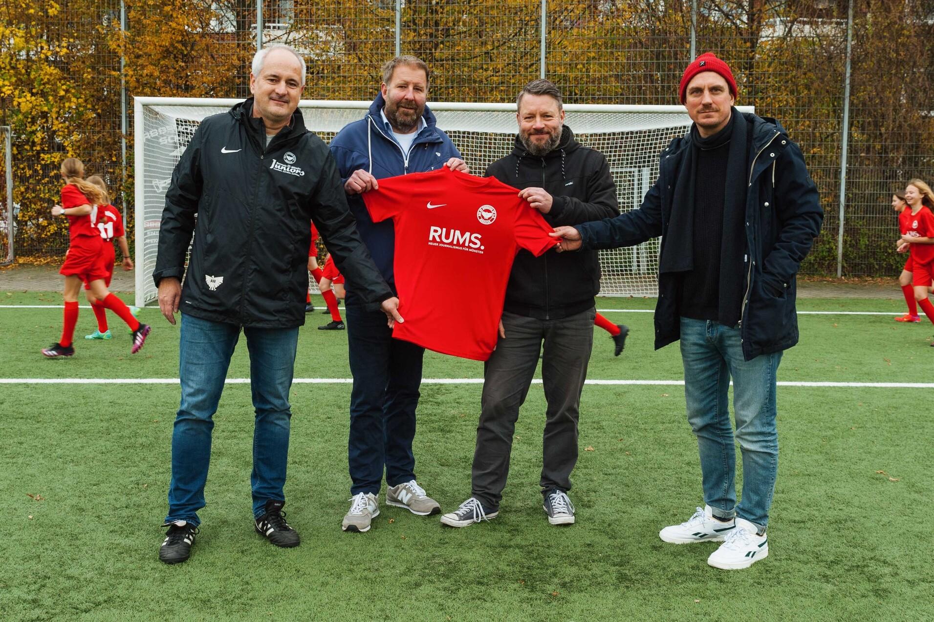 Vier M&auml;nner stehen auf einem Fu&szlig;ballplatz und halten ein rotes Trikot mit der Aufschrift &bdquo;RUMS&ldquo;. Im Hintergrund trainieren Kinder.