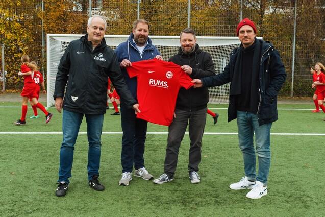 Vier M&auml;nner stehen auf einem Fu&szlig;ballplatz und halten ein rotes Trikot mit der Aufschrift &bdquo;RUMS&ldquo;. Im Hintergrund trainieren Kinder.