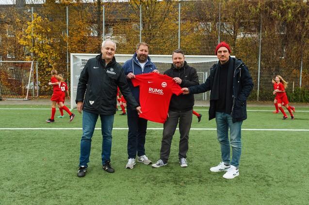 Vier M&auml;nner stehen auf einem Fu&szlig;ballplatz und halten ein rotes Trikot mit der Aufschrift &bdquo;RUMS&ldquo;. Im Hintergrund trainieren Kinder.