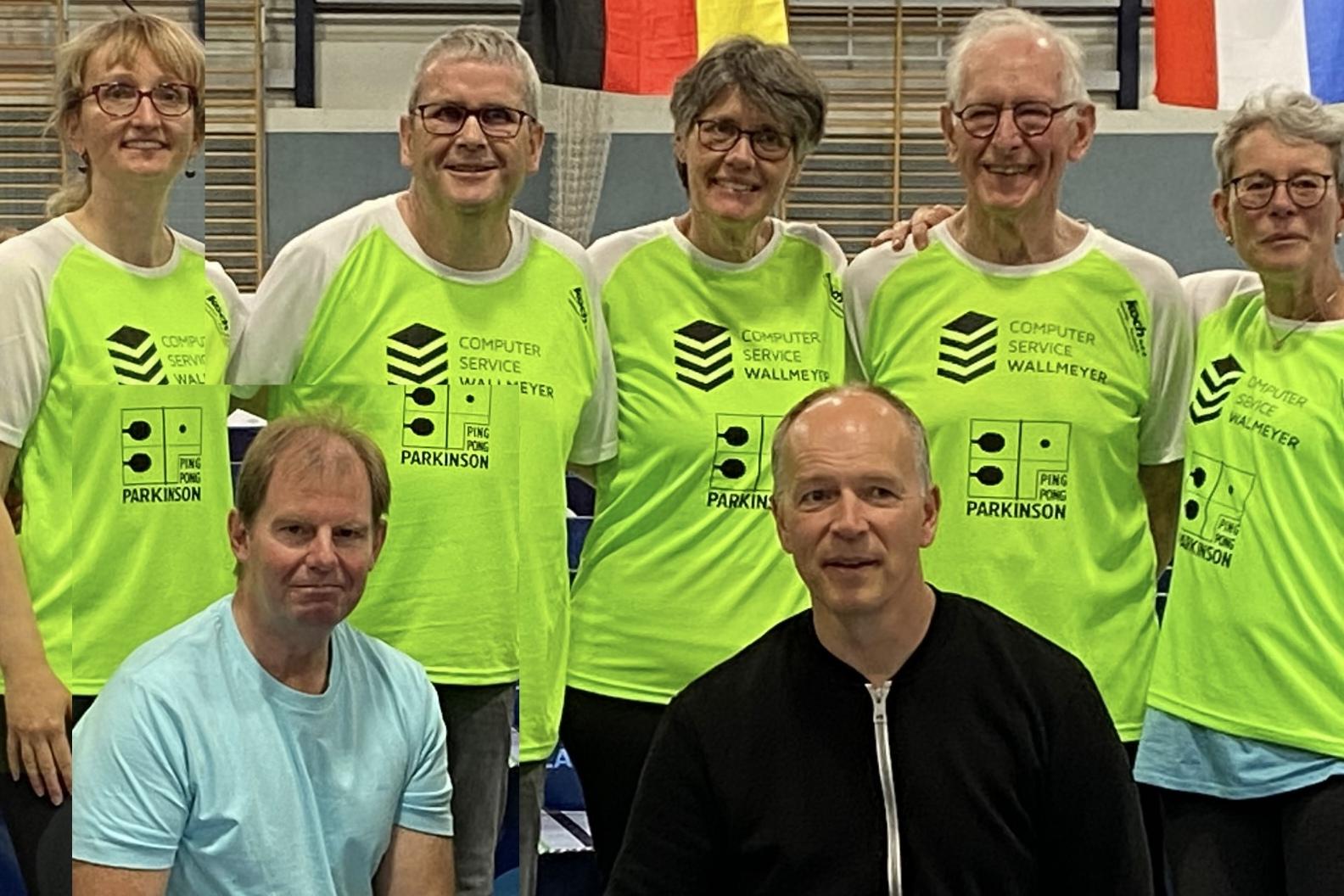Gruppe von sieben Personen in leuchtend gr&uuml;nen T-Shirts, posierend in einer Sporthalle mit Tischtennisausr&uuml;stung.