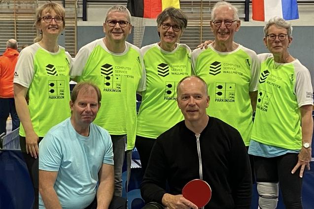 Gruppe von sieben Personen in leuchtend gr&uuml;nen T-Shirts, posierend in einer Sporthalle mit Tischtennisausr&uuml;stung.