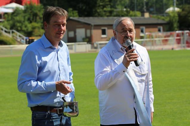 Zwei M&auml;nner stehen auf einem Fu&szlig;ballfeld, einer spricht ins Mikrofon, der andere h&auml;lt ein Glas in der Hand.