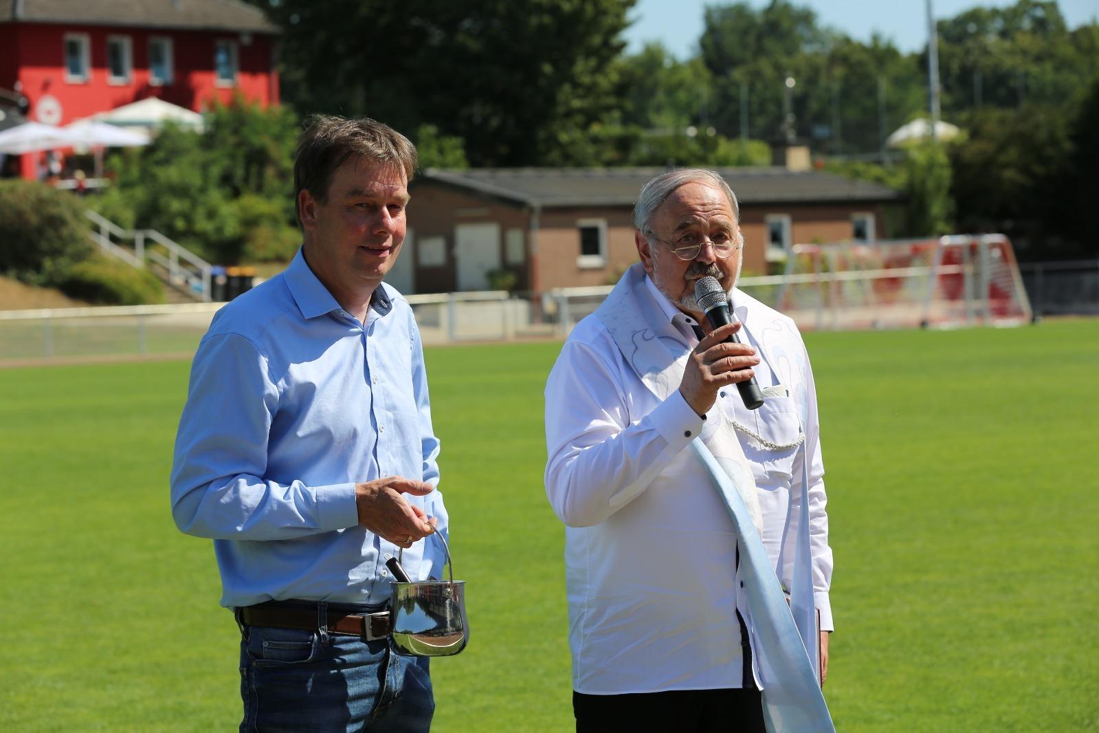Zwei M&auml;nner stehen auf einem Fu&szlig;ballfeld, einer spricht ins Mikrofon, der andere h&auml;lt ein Glas in der Hand.