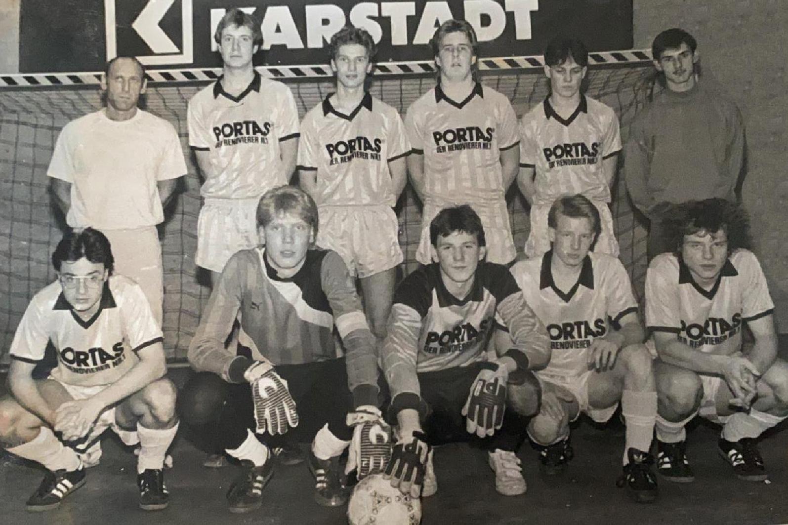 Schwarz-wei&szlig; Foto einer Fu&szlig;ballmannschaft mit Trainer, aufgestellt vor einem Tor mit &bdquo;Karstadt&ldquo;-Banner im Hintergrund.