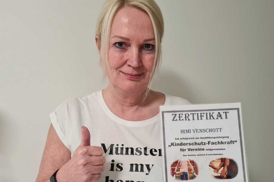 Frau mit Daumen hoch pr&auml;sentiert ein Zertifikat &uuml;ber "Kinderschutz-Fachkraft" vor einer wei&szlig;en Wand.