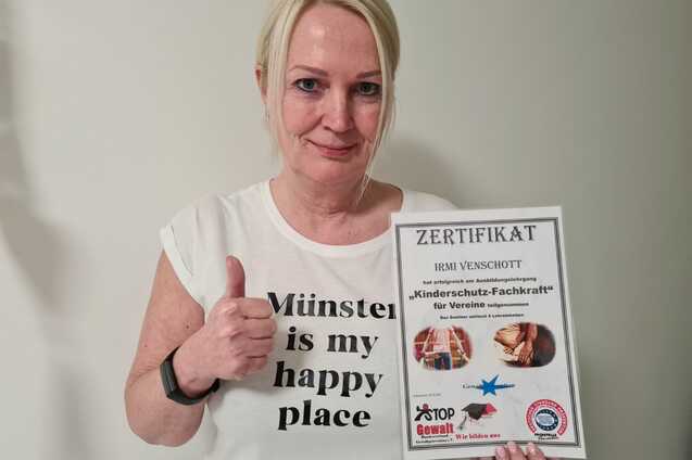 Frau mit Daumen hoch pr&auml;sentiert ein Zertifikat &uuml;ber "Kinderschutz-Fachkraft" vor einer wei&szlig;en Wand.