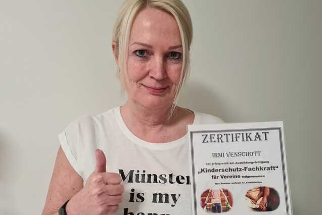 Frau mit Daumen hoch pr&auml;sentiert ein Zertifikat &uuml;ber "Kinderschutz-Fachkraft" vor einer wei&szlig;en Wand.