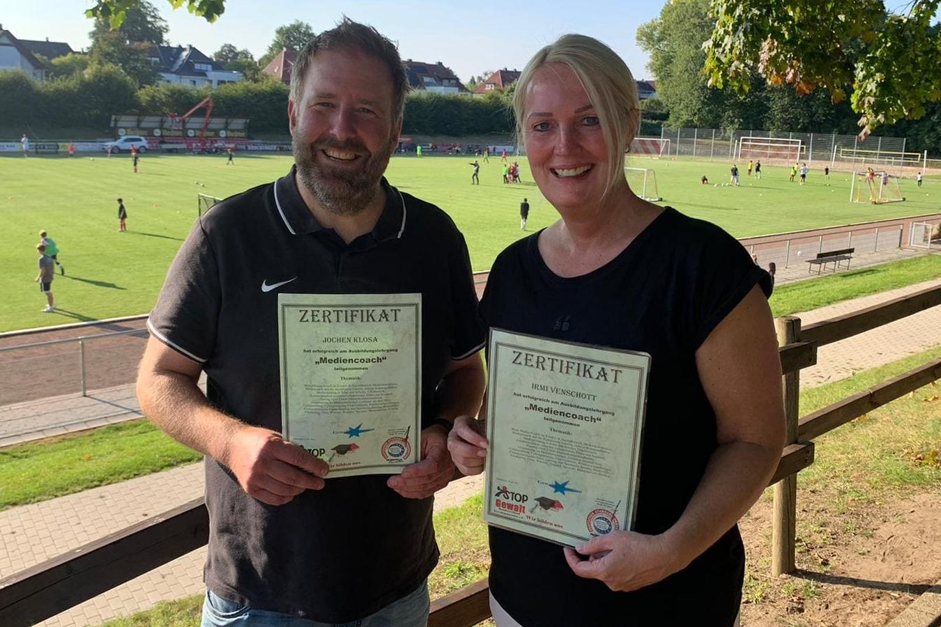 Zwei Personen halten stolz ihre Zertifikate vor einem Sportplatz mit Fu&szlig;ballspielern im Hintergrund.