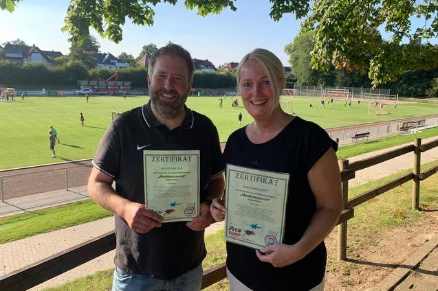 Zwei Personen halten stolz ihre Zertifikate vor einem Sportplatz mit Fu&szlig;ballspielern im Hintergrund.
