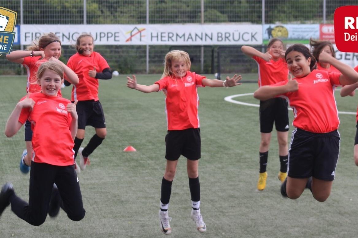 Sechs M&auml;dchen in roten Trikots springen fr&ouml;hlich auf einem Fu&szlig;ballfeld, umgeben von H&uuml;tchen und Werbebannern.