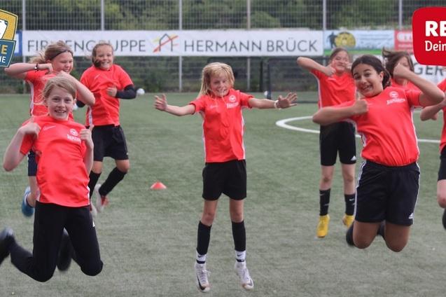 Sechs M&auml;dchen in roten Trikots springen fr&ouml;hlich auf einem Fu&szlig;ballfeld, umgeben von H&uuml;tchen und Werbebannern.