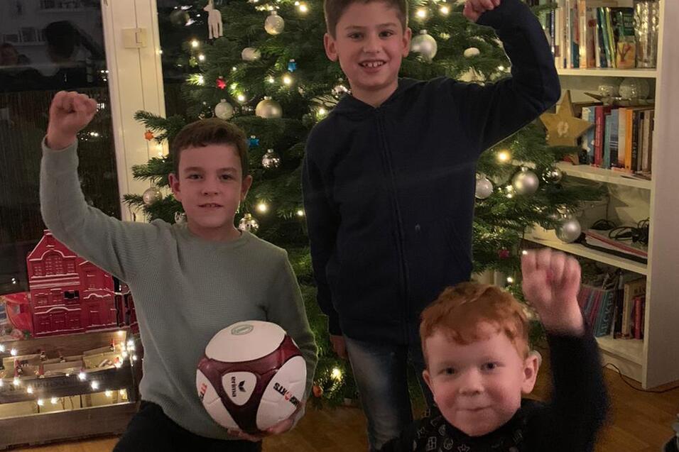 Drei Kinder stehen vor einem beleuchteten Weihnachtsbaum und halten jeweils einen Fu&szlig;ball in den H&auml;nden.