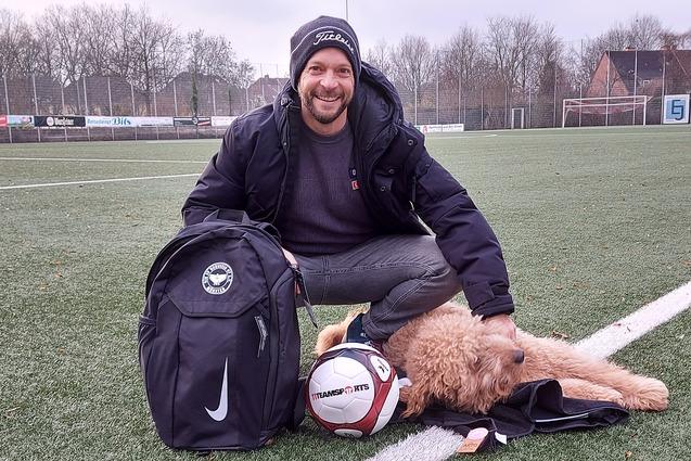L&auml;chelnder Mann mit Rucksack sitzt auf einem Fu&szlig;ballfeld neben einem Hund und einem Fu&szlig;ball.