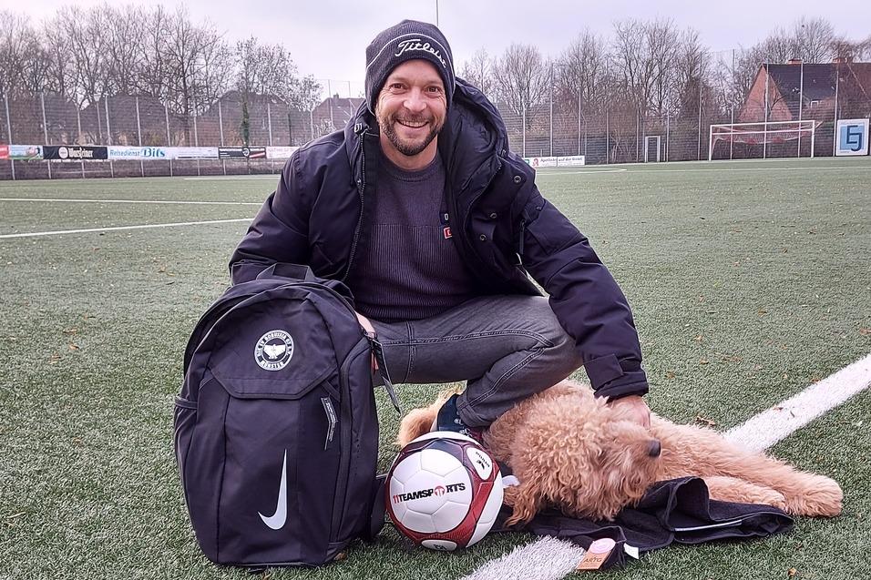 L&auml;chelnder Mann mit Rucksack sitzt auf einem Fu&szlig;ballfeld neben einem Hund und einem Fu&szlig;ball.