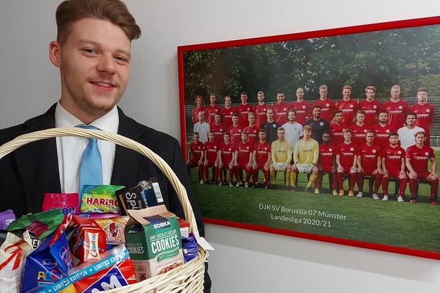 Mann in Anzug h&auml;lt einen geflochtenen Korb voller Snacks vor einem Mannschaftsbild des DJK SV Borussia 07 M&uuml;nster.