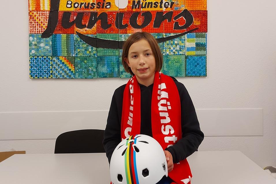 Ein M&auml;dchen mit kurzgeschnittenen Haaren sitzt an einem Tisch, h&auml;lt einen Fahrradhelm und tr&auml;gt einen Schal von Borussia M&uuml;nster.