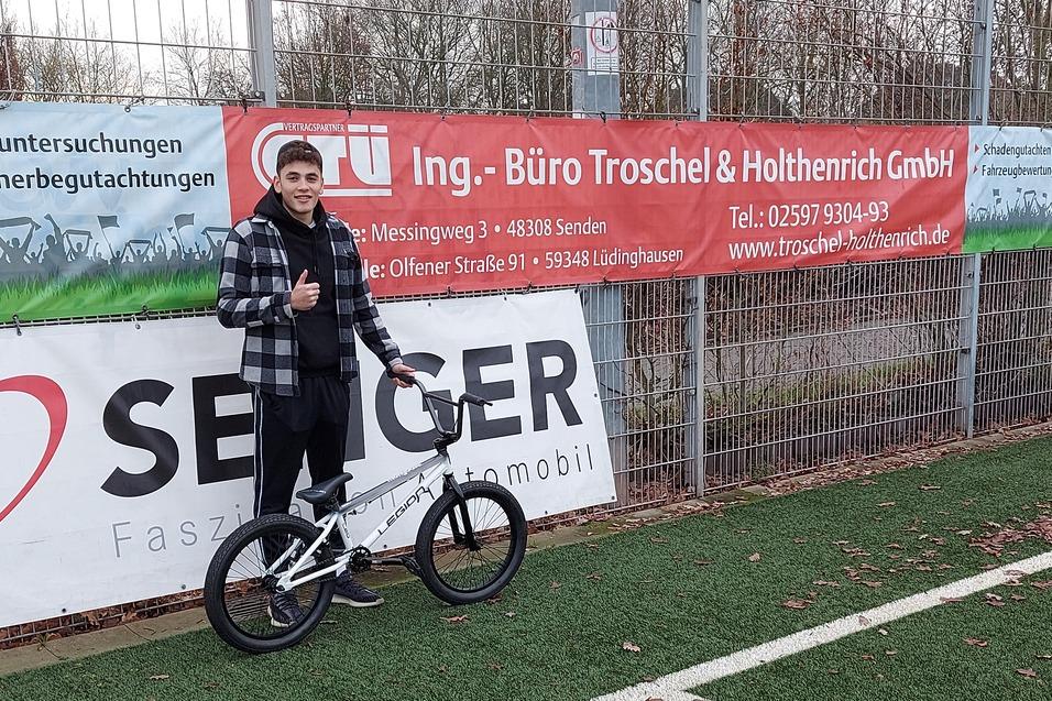 Junger Mann mit BMX-Fahrrad steht auf einem Kunstrasenplatz und zeigt Daumen hoch, Werbebanner im Hintergrund.