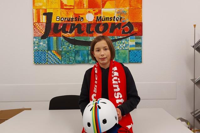 Ein M&auml;dchen mit kurzgeschnittenen Haaren sitzt an einem Tisch, h&auml;lt einen Fahrradhelm und tr&auml;gt einen Schal von Borussia M&uuml;nster.