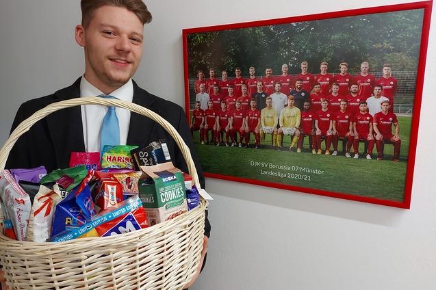 Mann in Anzug h&auml;lt einen geflochtenen Korb voller Snacks vor einem Mannschaftsbild des DJK SV Borussia 07 M&uuml;nster.