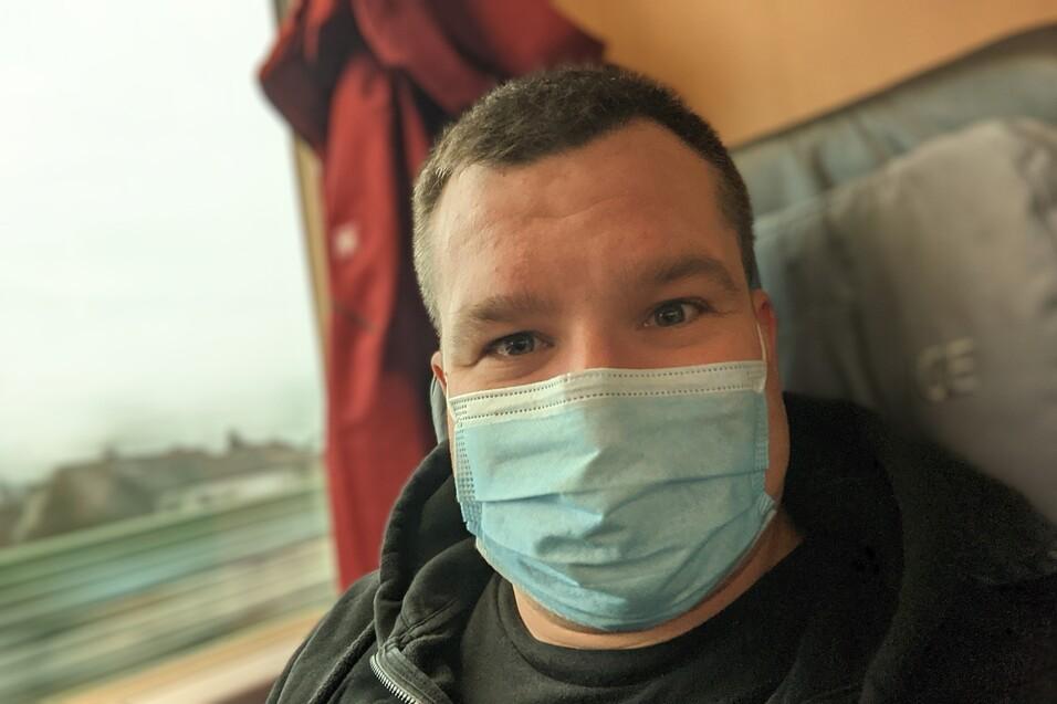 Mann mit Mundschutz sitzt im Zug und l&auml;chelt, Hintergrund zeigt verschwommene Landschaft.