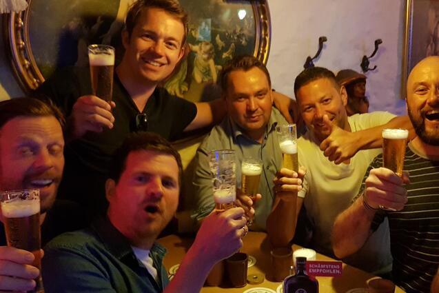 Gruppierung von sieben M&auml;nnern, die an einem Tisch in einer Bar Biergl&auml;ser heben und l&auml;cheln, umgeben von W&auml;nden und Dekor.