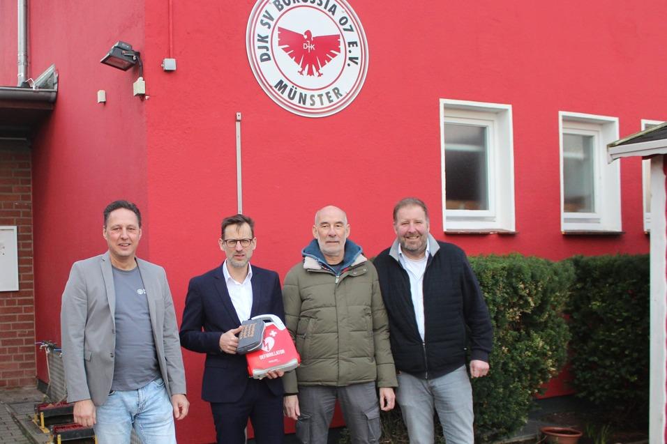 Vier M&auml;nner stehen vor einem roten Geb&auml;ude mit dem Logo von Borussia M&uuml;nster, w&auml;hrend sie Fahnen in den H&auml;nden halten.