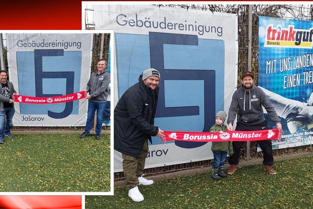 Drei Personen, darunter ein Kind, halten ein Fan-Banner von Borussia M&uuml;nster vor einem Werbebanner.