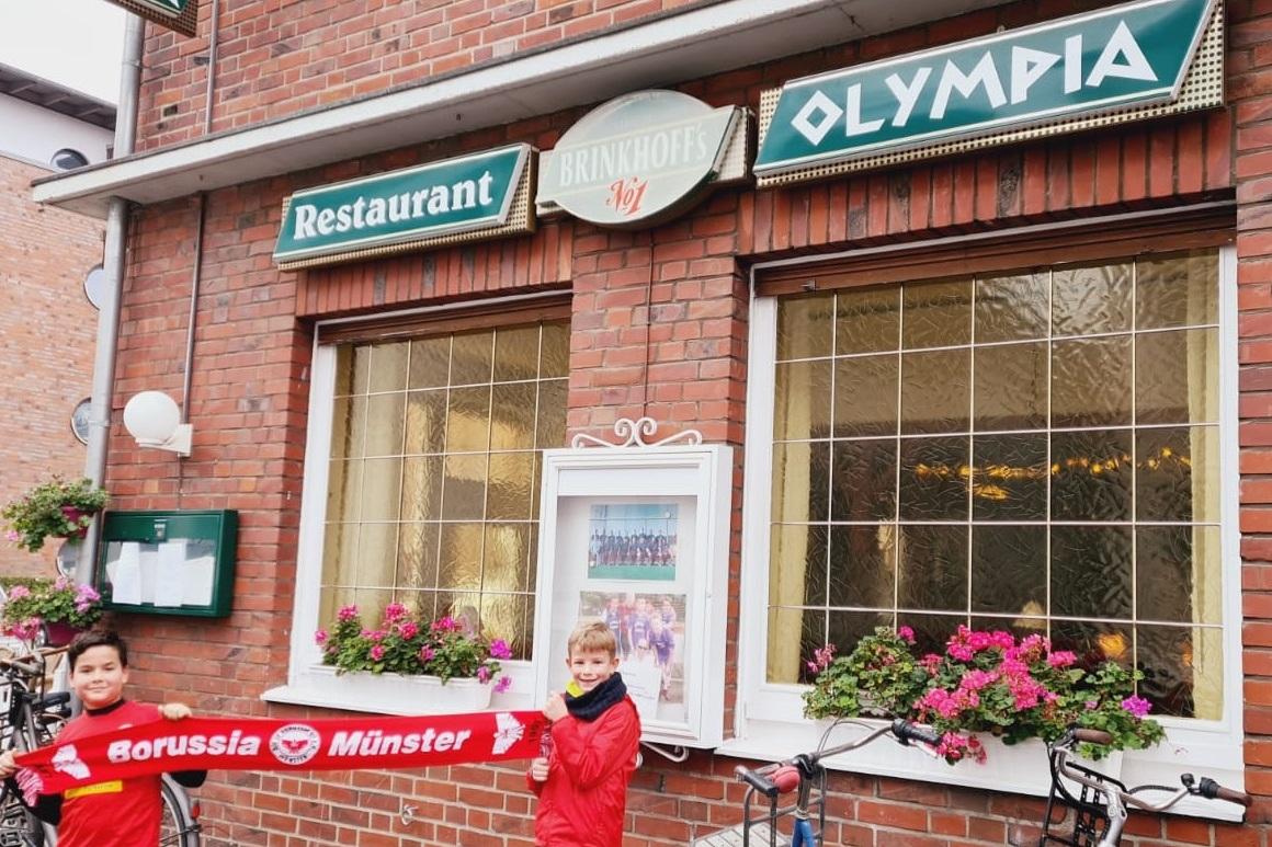 Zwei Kinder in roten Trikots halten ein Banner von Borussia M&uuml;nster vor einem Restaurant mit Blumenfenstern.