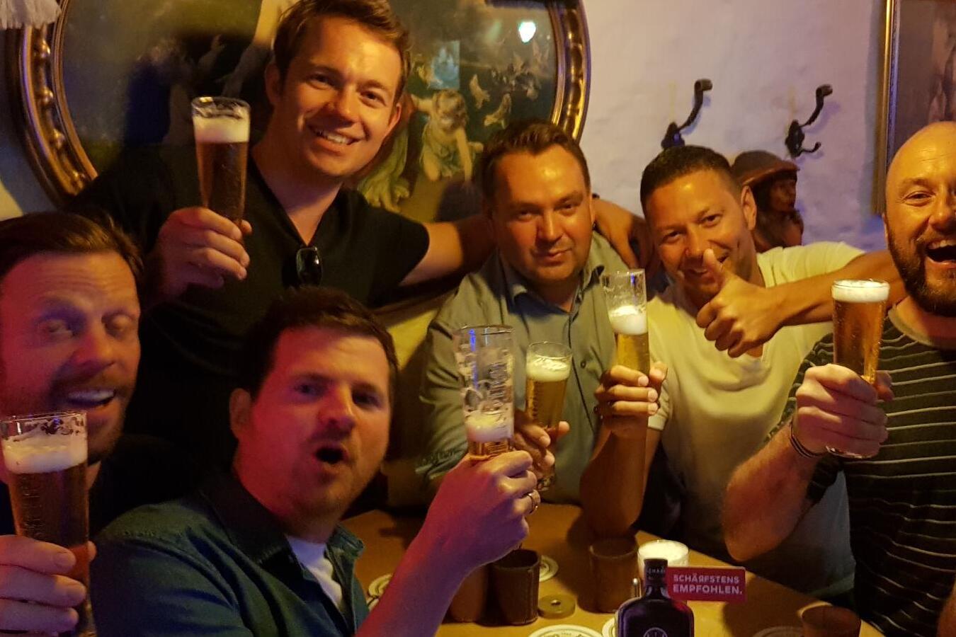 Gruppierung von sieben M&auml;nnern, die an einem Tisch in einer Bar Biergl&auml;ser heben und l&auml;cheln, umgeben von W&auml;nden und Dekor.