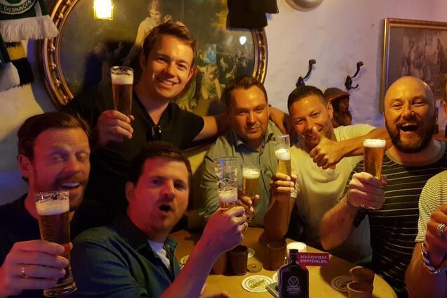 Gruppierung von sieben M&auml;nnern, die an einem Tisch in einer Bar Biergl&auml;ser heben und l&auml;cheln, umgeben von W&auml;nden und Dekor.