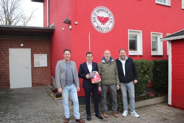 Vier M&auml;nner stehen vor einem roten Geb&auml;ude mit dem Logo von Borussia M&uuml;nster, w&auml;hrend sie Fahnen in den H&auml;nden halten.