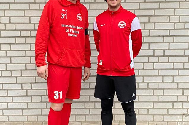 Zwei Fu&szlig;ballspieler in roter Sportkleidung stehen vor einer gemusterten Backsteinwand.
