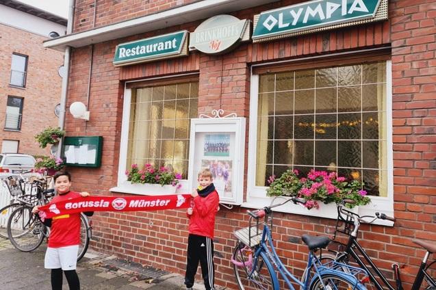 Zwei Kinder in roten Trikots halten ein Banner von Borussia M&uuml;nster vor einem Restaurant mit Blumenfenstern.