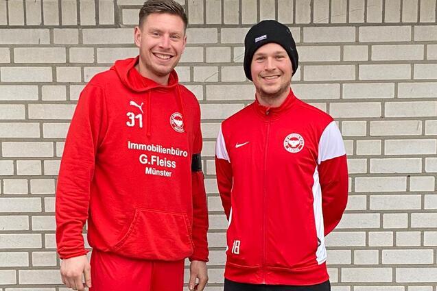 Zwei Fu&szlig;ballspieler in roter Sportkleidung stehen vor einer gemusterten Backsteinwand.