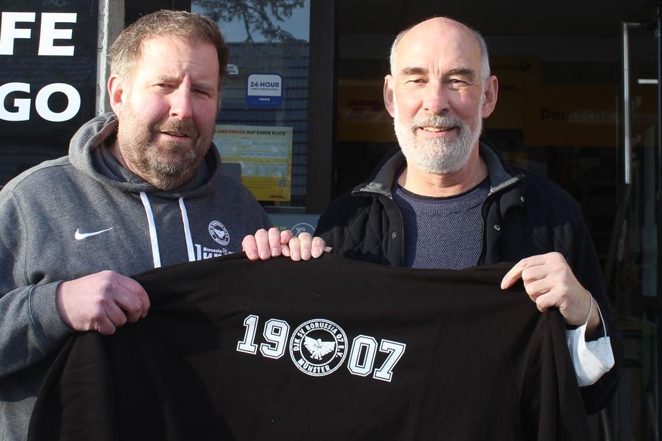 Zwei M&auml;nner stehen vor einem Caf&eacute; und halten ein schwarzes T-Shirt mit der Aufschrift &bdquo;19007&ldquo;. Im Hintergrund ist das Caf&eacute;-Schild sichtbar.