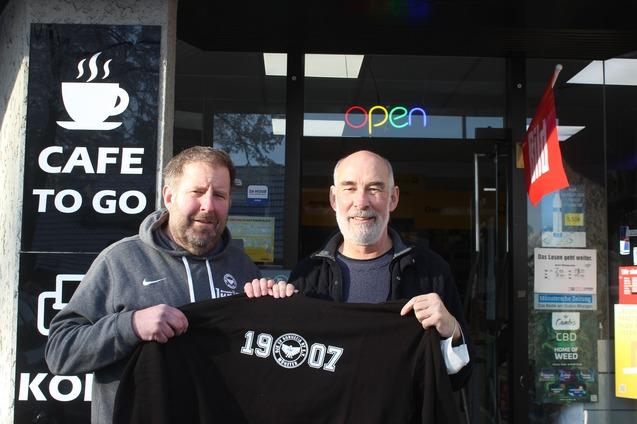Zwei M&auml;nner stehen vor einem Caf&eacute; und halten ein schwarzes T-Shirt mit der Aufschrift &bdquo;19007&ldquo;. Im Hintergrund ist das Caf&eacute;-Schild sichtbar.