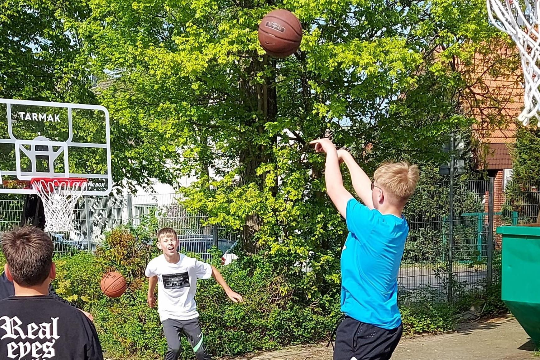 Junger Spieler wirft Basketball zum Korb, w&auml;hrend andere Spieler im Hintergrund warten, auf einem Freiplatz.