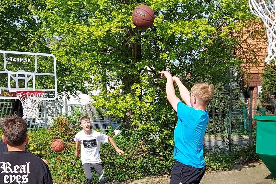 Junger Spieler wirft Basketball zum Korb, w&auml;hrend andere Spieler im Hintergrund warten, auf einem Freiplatz.