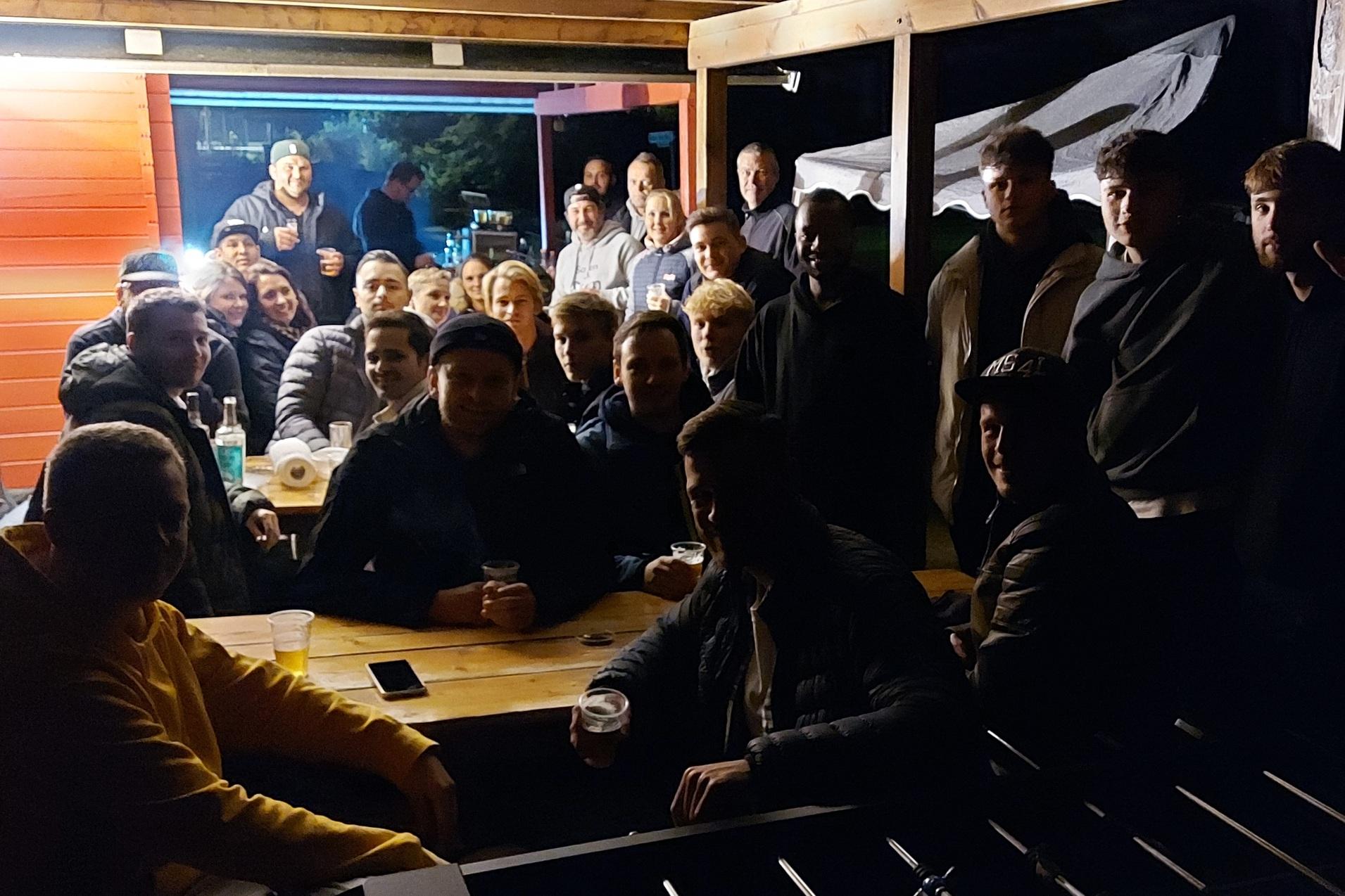 Gruppe von M&auml;nnern sitzt an einem Tisch unter einem &uuml;berdachten Bereich bei Nacht, mit Getr&auml;nken und einer warmen Atmosph&auml;re.