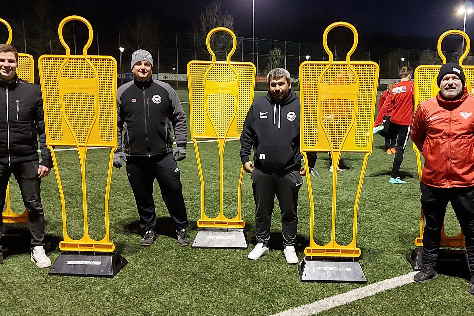 Training auf dem Fu&szlig;ballplatz mit vier orangefarbenen Trainingshilfen und drei Trainern in Sportkleidung.